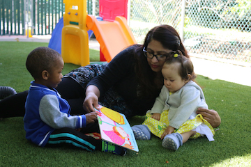 A teacher and two infants look at a book outdoors
