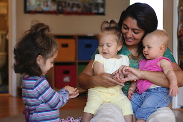 Teacher with two infants on her lap interacting with a toddler who is standing next to them